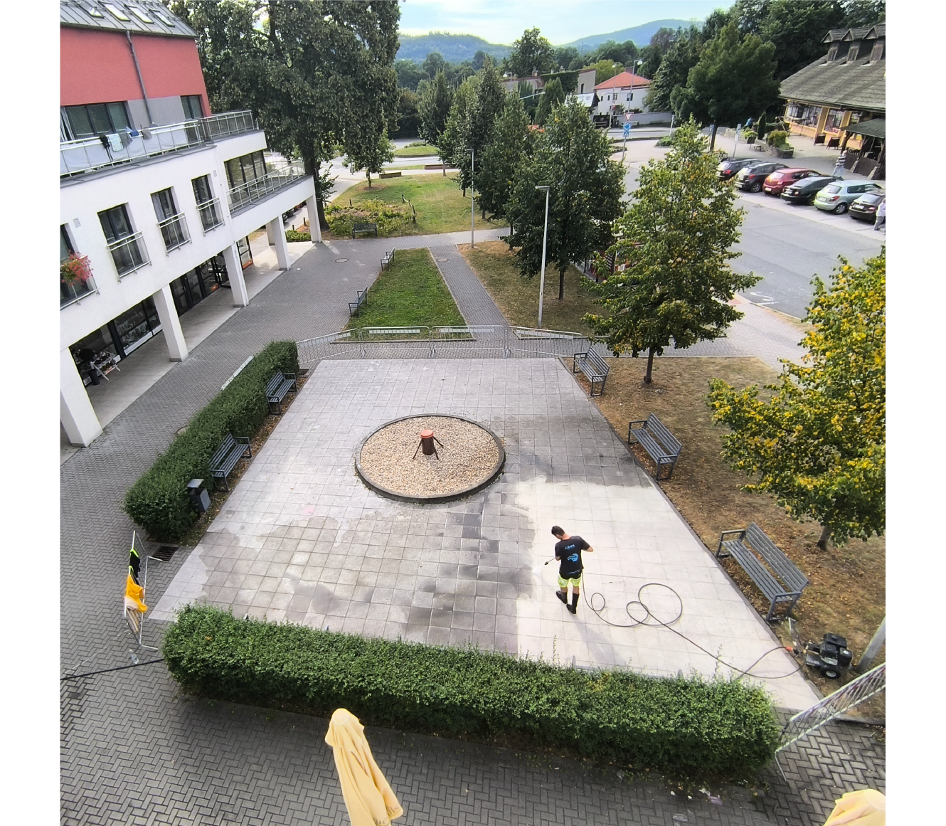 Person pressure washing a large tiled courtyard with benches, trees, and surrounding buildings visible.